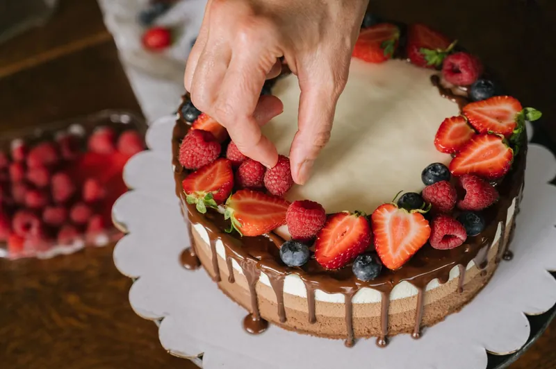 Hands placing fresh strawberries and blueberries on a chocolate-drip cake